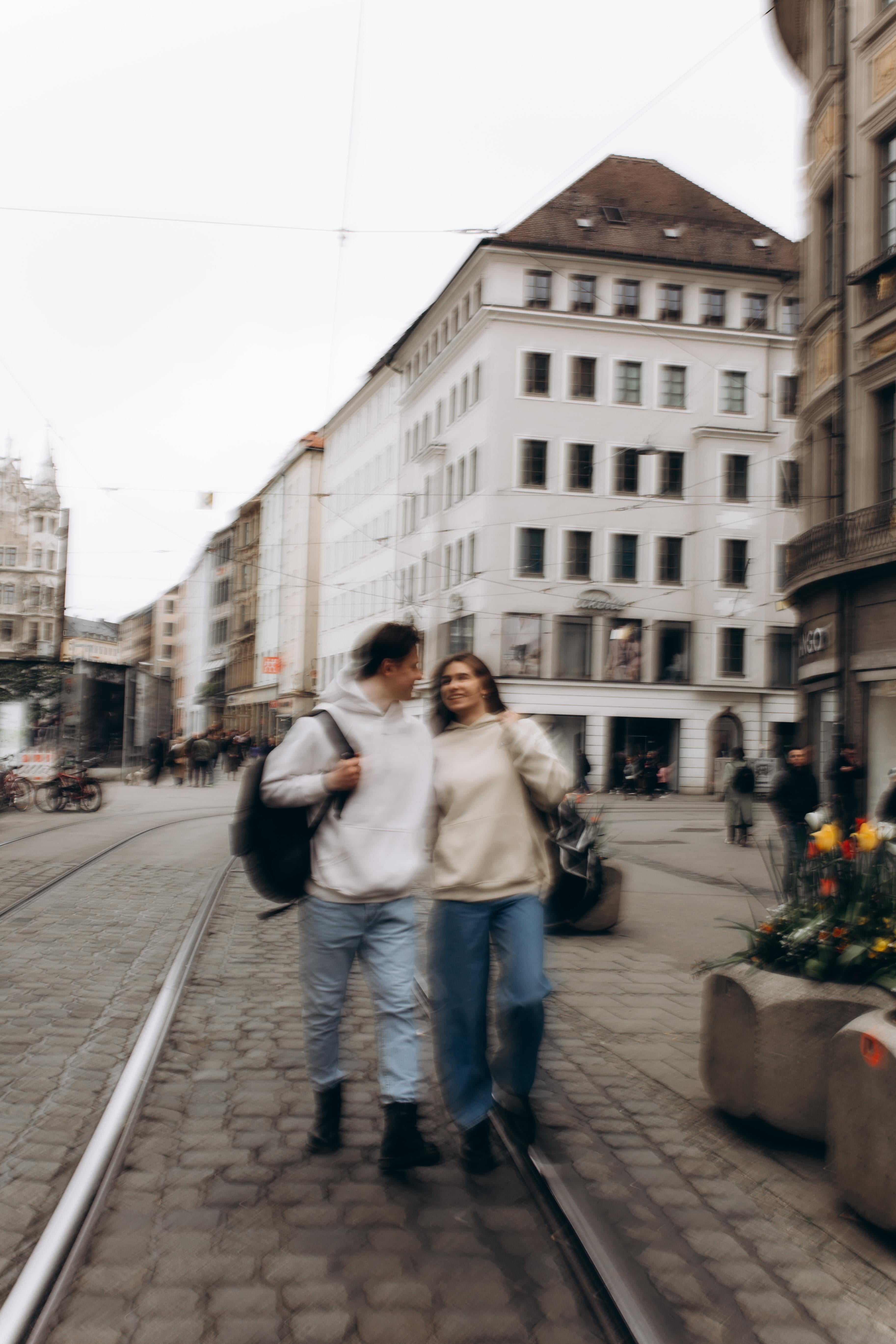 Two people walking on a city street with buildings and a tram track in the background.